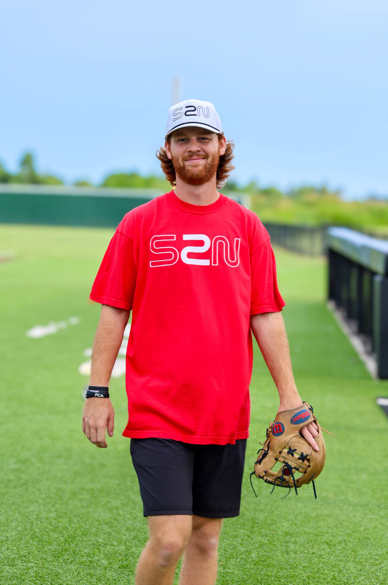 Man in a red shirt with 'S2N' logo holding a baseball glove on a sports field.